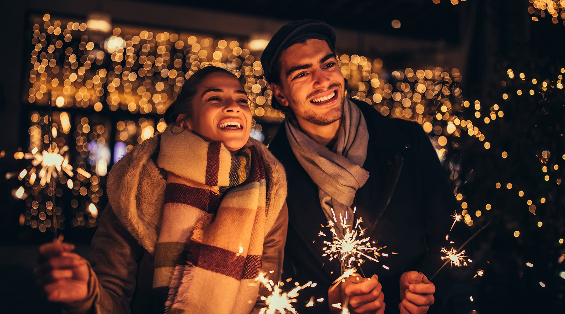 Smiling couple at nighttime dressed warmly holding sparklers with sparkling lights behind them